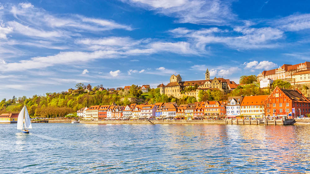 Panoramic view on the charming medieval city of Meersburg, located at the Lake Constance (Bodensee), with a sailing boat passing the city´s beautiful waterfront in the lower town and it´s landmark castles in the upper town. 
