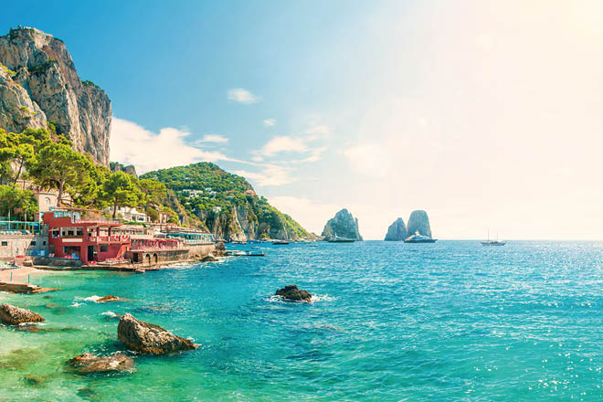 view of faraglioni rocks at sunset from Marina Piccola beach on summer day with turquoise blue waters and beachfront cafes, Capri, Italy