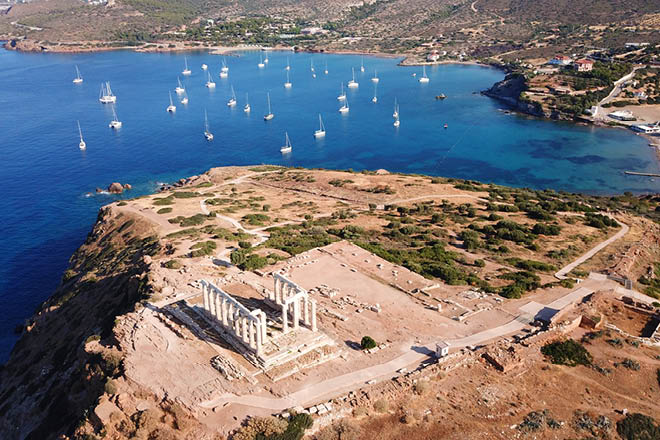 Aerial drone bird's eye view of archaeological site of Cape Sounio and magnificent deep blue bay with yachts docked, Ancient temple of Poseidon, Attica, Greece