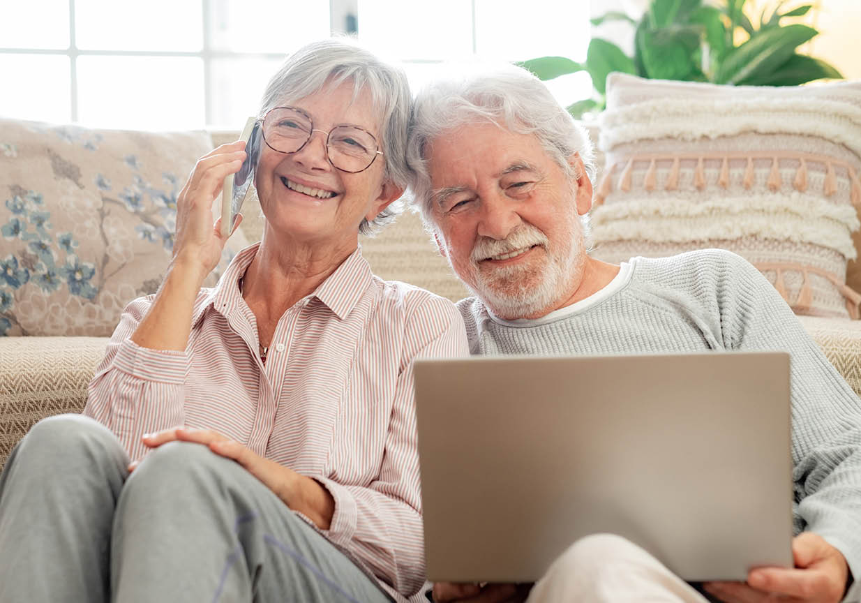 Beautiful senior white-haired couple sitting on floor at home, husband browsing laptop wife talking on phone. Concept of family and serene retirement