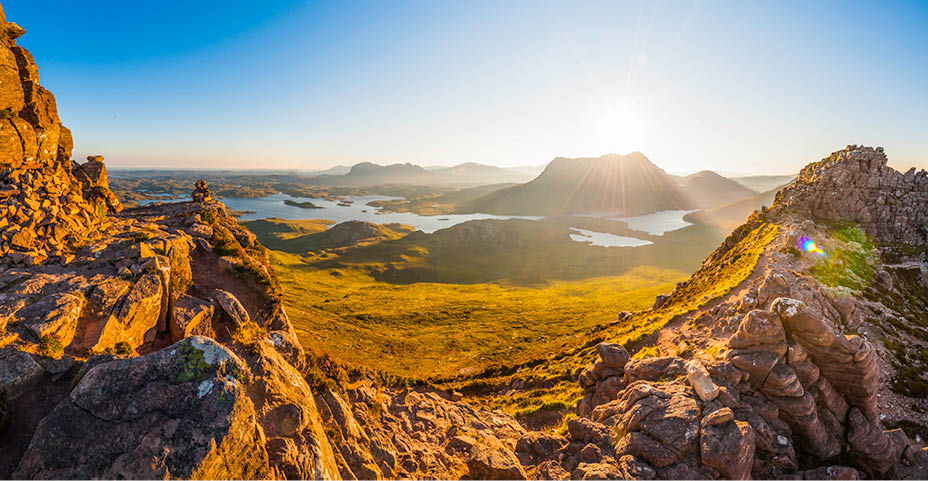 Warm early morning sunlight bursting over the mountain peaks of Cul Mor, Suilven and Canisp to the dramatic wilderness landscape of Inverpolly and Assynt illuminating the golden rocks and heather of Stac Pollaidh, deep in the Sutherland Highlands of Scotland.